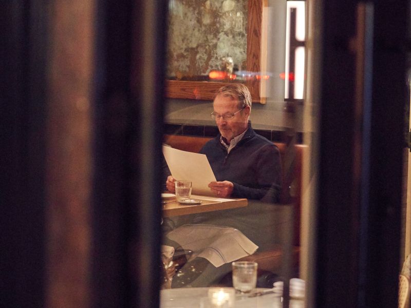 A guest reading the menu in a leather booth at St. Neo's Brasserie, seen through the window